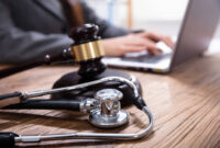 Close-up Of Mallet And Stethoscope On Gavel Over The Office Desk With Businessperson Using Laptop